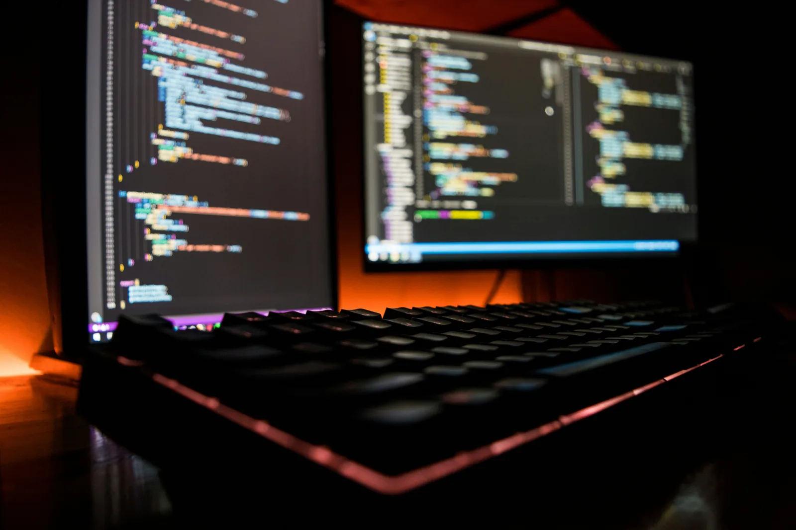 Zwei Bildschirme mit farbig hervorgehobenem Quellcode hinter einer Tastatur, in orangefarbenem Licht